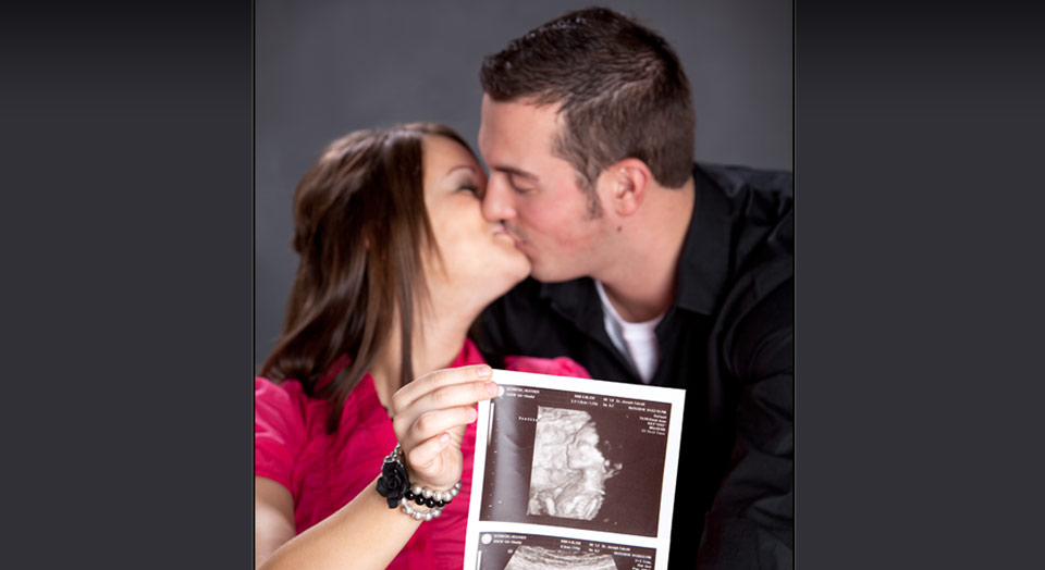 Maternity photography of couple holding out ultrasound picture while kissing